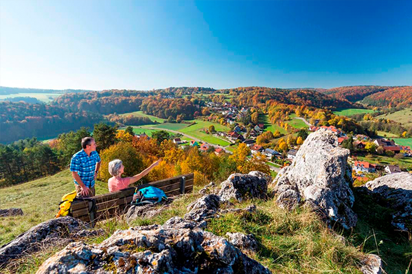 Imagebild Wandern: Pärchen genießt die Aussicht über die Landschaft von einer Holzbank aus